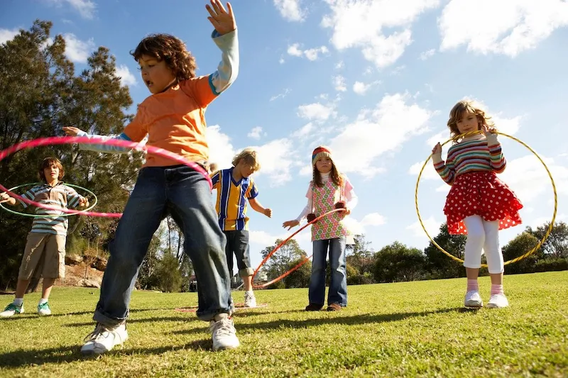 Hoola Hooping featured image Children joyfully hula hooping on a sunny day in a grassy park, with trees and a blue sky in the background. The scene is lively and playful.