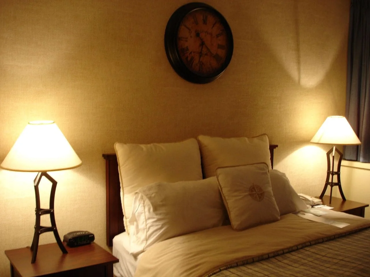Sleep Strategies in Times of Stress featured image Cozy bedroom with a warm, inviting ambiance. A neatly made bed features multiple pillows, flanked by lit table lamps. A vintage clock hangs above.