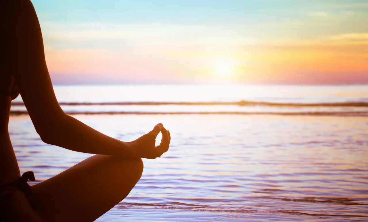 just breathe featured image A person meditates on a beach at sunrise, sitting cross-legged with one hand in a mudra pose. The tranquil scene features a colorful sky and calm ocean waves.