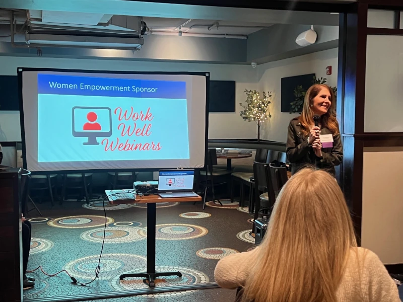 A woman speaks at a podium in a cozy room, smiling, under soft lighting. Behind her, a screen displays "Women Empowerment Sponsor: Work Well Webinars."